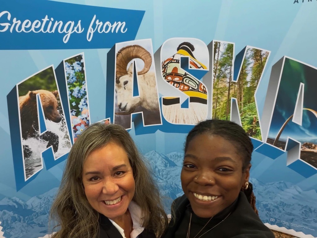 Two students smiling together in front of an Alaska-themed backdrop featuring wildlife, forests, and scenic landscapes during their Alaska trip.